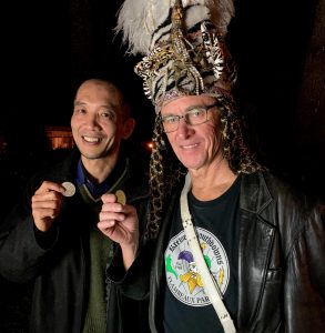 Two men face the camera: at left, Naohiro Kato, and at right, a Mardi Gras krewe leader wearing a headdress. Both hold small bioplastic doubloons.